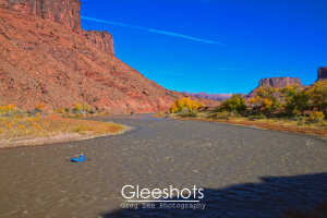 A  small blue raft floats down the Colorado River in Moab, surrounded by red rock cliffs and autumn foliage under a clear blue sky.   Utah Fine Art Landscape Photography Print for Sale.