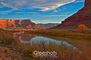 The winding Colorado River reflects red rock canyon walls and a blue sky with wispy clouds at sunset. Autumn foliage adds color to the landscape, with the Fisher Towers and  snow-capped mountains in the distance.  Utah Fine Art Landscape Photography for Sale.