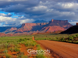 In Moab, a dirt road leads through a green, open plain towards striking red rock mesas beneath a dramatic, cloudy sky. The snow-capped La Sal mountains are visible in the background.   Utah Fine Art Landscape Photography for Sale.