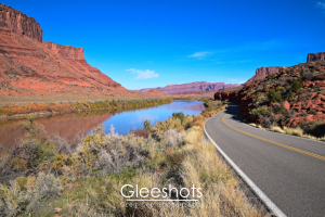 The winding road of Scenic Byway 128 runs beside the Colorado River in Moab, surrounded by red rock cliffs and desert vegetation under a bright blue sky.  Utah Fine Art Landscape Photography Print for Sale.