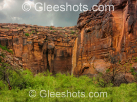 A dramatic red rock cliff formation resembling a face rises above green shrubs under storm clouds in Moab.  Rain from the storm has created a waterfall, resembling tears from the eye of the face.  Utah Fine Art Landscape Photography for Sale.