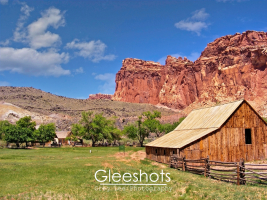 In Capitol Reef National Park, the rustic wooden Pendleton Barn and split-rail fence sit in a green pasture, with scattered trees and dramatic red rock cliffs in the background.  National Park Landscape Photography for Sale.