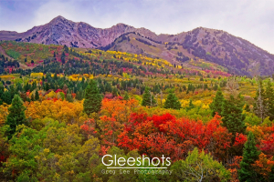 Sun shines on the mountains near Snowbasin Utah,  highlighting colorful autumn trees in shades of red, orange, yellow, and green, with rugged mountains rising in the background under a cloudy sky.  Fine Art Landscape Photography for Sale.