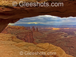 A view of Canyonlands National Park through Mesa Arch.  Layers of red rock formations stretch into the distance under a cloudy sky, with beams of filtered sunlight shining on the canyon floor in the distance.  Utah Fine Art Landscape Photography for Sale.