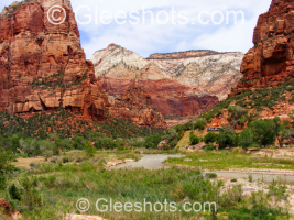 A panoramic view of Zion National Park with dramatic red rock cliffs, green vegetation, the winding Virgin river, and a shuttle bus traveling along the road.  National Park Landscape Photography Print for Sale.