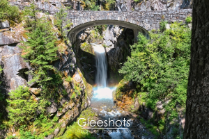 A stone bridge arches over Christine Falls, with sunlight creating a faint rainbow in the mist below.  The waterfall cascades into a gorge surrounded by lush green trees.  Rainier Landscape Photography for Sale.