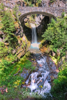 Christine Falls in Mt Rainier National Park flows beneath a stone arch bridge surrounded by lush green vegetation.  A rainbow is visible as the waterfall cascades into the stream below.  Washington Fine Art Landscape Photography for Sale.