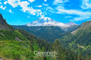 A snow-capped Mount Rainier looms over a lush, green valley with a mix of pine forests and blue skies spotted with clouds in this view from Stevens Canyon Road.  Washington Landscape Photography Print for Sale.
